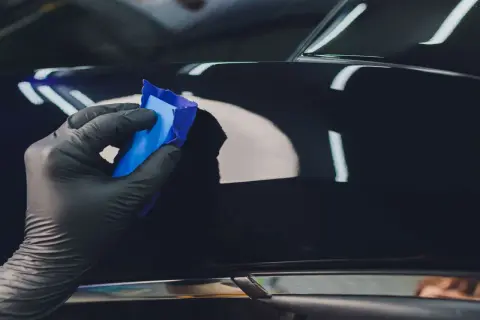 Ręka w czarnej rękawiczce nakłada niebieską glinkę na błyszczącą karoserię, wykonując serwis powłoki ceramicznej.