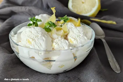 Ein Glas cremiges zitronen eis, garniert mit Minze und Zitronenschale, daneben eine halbe Zitrone und ein Löffel.