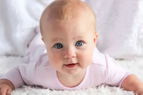 Bebé con ojos azules y cabello rubio, con un mameluco rosa. Pensando en nombres de chica ingleses como Emily o Olivia.