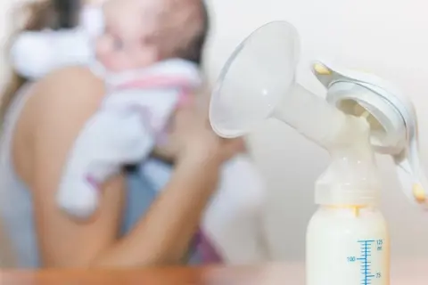 Laktator z butelką mleka, w tle mama z dzieckiem. Nawał pokarmu w której dobie to wyzwanie, ale dzięki niemu maluch ma co jeść.