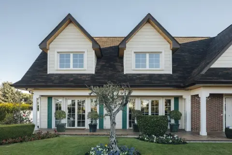 Fachada de casas Canexel con ventanas blancas, contraventanas verdes y un jardín cuidado con un olivo.