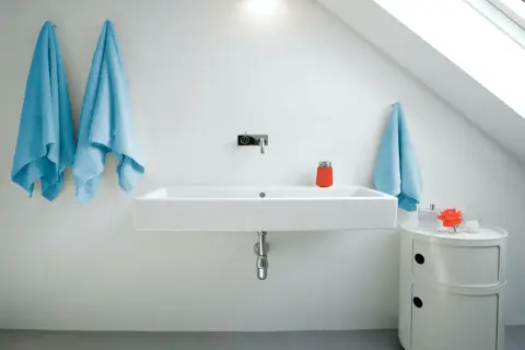 Modernt badrum med ett stilrent handfat. Två ljusblåa handdukar hänger på vänster sida, en på höger. En orange behållare och en blomma står på ett litet vitt skåp.
