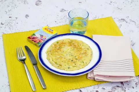 Tortilla de gambas dorada en plato blanco con borde azul, servida con cubiertos, vaso de agua y caldo de pescado.