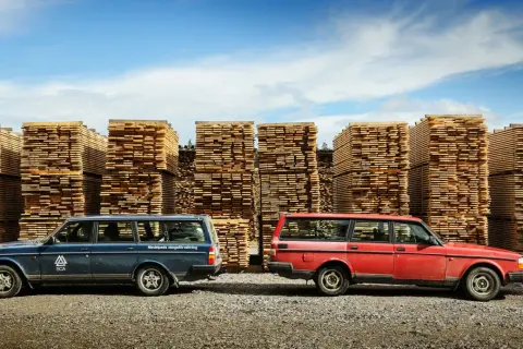 Två Volvo 240 kombi, en blå och en röd, står framför höga staplar av träplankor under en blå himmel.
