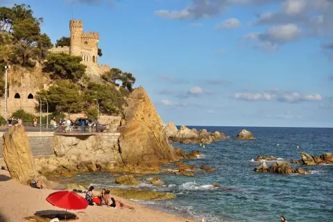 El kasteel Lloret de Mar se alza sobre una colina rocosa junto a una playa con gente disfrutando del sol y el mar azul.