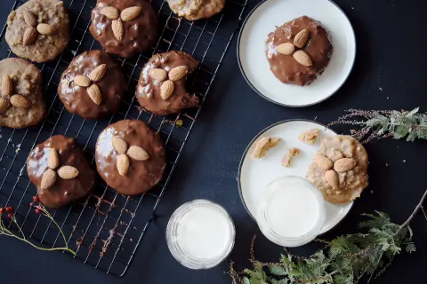 Schokoladenüberzogene Elisenlebkuchen mit Marzipan und Mandeln auf einem Gitter, daneben Milchgläser und Tannenzweige.