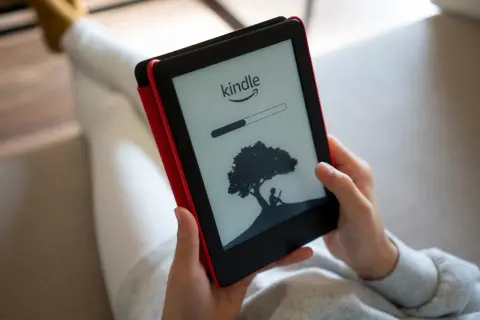 Une personne lit sur une liseuse Kindle, affichant une barre de progression et une silhouette sous un arbre. Un guide d'utilisation Kindle est à portée de main.
