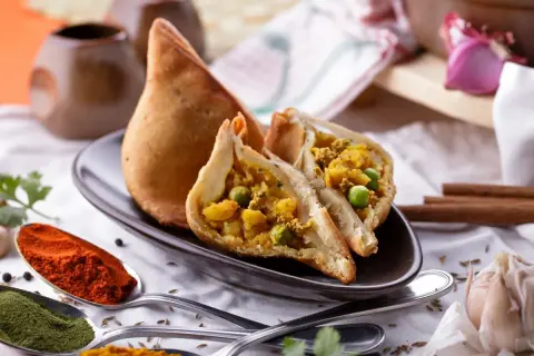Chrupiący indyjski pierożek samosa, przekrojony na pół, ukazuje bogate nadzienie z ziemniaków i groszku. Obok łyżki z papryką i zielonymi przyprawami.