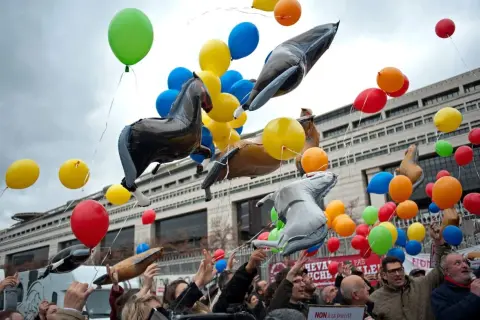 Un lâcher de ballons colorés et gonflables en forme de chevaux s'envole devant un bâtiment imposant.