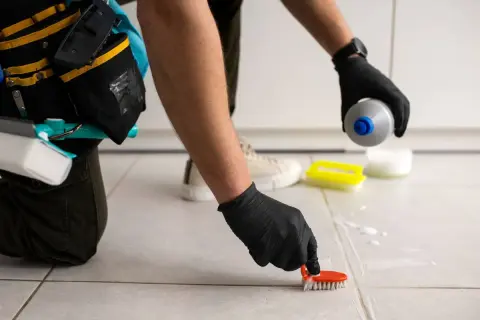 Persona con guantes negros usa un cepillo para limpiar juntas de azulejos, aplicando producto de limpieza.