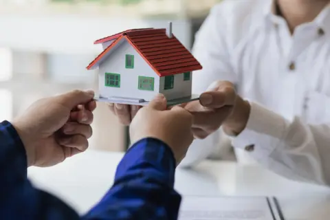 Manos intercambian una maqueta de casa, simbolizando la decisión de vender herencia.