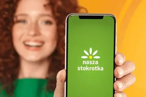 Uśmiechnięta kobieta prezentuje ekran telefonu z zieloną aplikacją "Nasza Stokrotka".