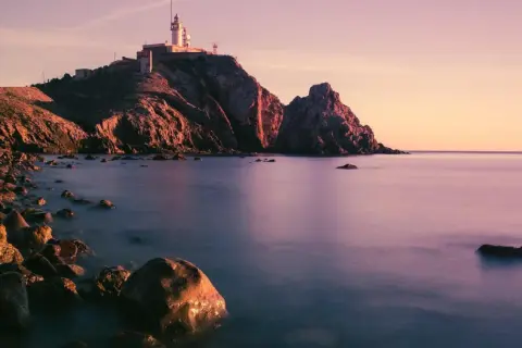 Faro en un acantilado rocoso al atardecer, con el mar tranquilo y piedras en la orilla. Parece la boca de los frailes.