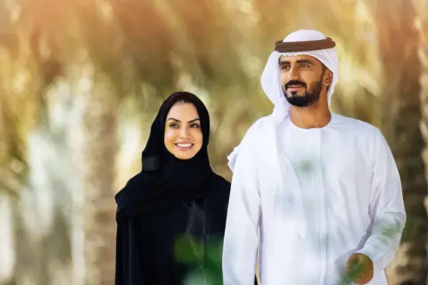Uśmiechnięta kobieta w tradycyjnym stroju arabskim damskim i mężczyzna w białej dżalabiji spacerują wśród palm.