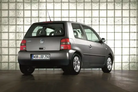 Szary Volkswagen Lupo stoi na ciemnej podłodze, na tle ściany z białych pustaków szklanych.