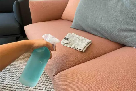 Hand sprüht Reinigungsmittel auf einen rosafarbenen Sofa-Stoff, um Wasserflecken zu entfernen. Ein Tuch liegt daneben.
