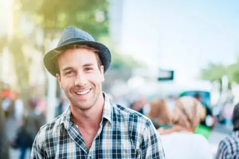 Uomo sorridente con un cappello adatto al viso, indossa una camicia a quadri. La luce del sole illumina la scena.