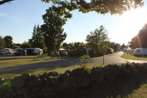 Solig dag på haga park camping. Husvagnar och bilar står uppradade på gräsmattor.