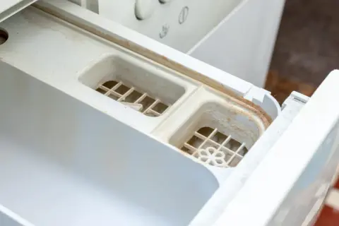 Schmutziges Waschmittelfach einer Waschmaschine, das dringend eine Reinigung benötigt. Kalk und Seifenreste haben sich abgelagert.