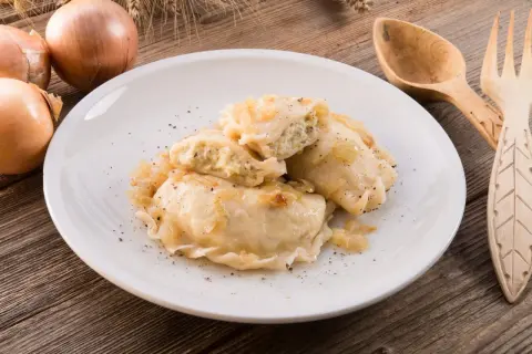 Pierogi z serem i cebulką na białym talerzu, obok cebule i drewniane sztućce.