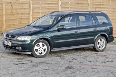 Zielony Opel Astra kombi, jeden z wielu aut w ocynku, stoi na mokrym parkingu przed drewnianym płotem.
