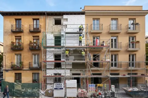 Trabajadores en andamios renuevan fachadas de edificios, aplicando aislamiento térmico. Se ve un cartel de "OBRA SATE".