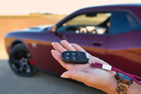 Kluczyk do nowego auta w dłoni. To pierwszy krok w kupnie samochodu krok po kroku. Czerwony Dodge Challenger w tle.