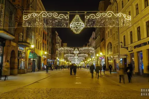 Toruń stare miasto nocą, udekorowane świątecznymi lampkami. Brukowana ulica pełna ludzi, otoczona kamienicami.