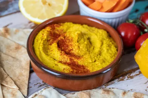En skål med krämig hummus, toppad med paprikapulver, serveras med pitabröd, citron och grönsaker. Perfekt för ett enkelt hummus recept.
