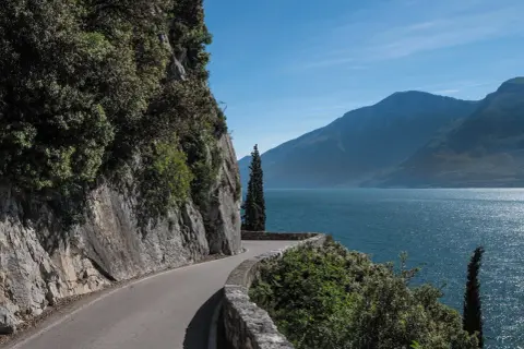 Strada panoramica lungo il lago, con rocce a sinistra e montagne sullo sfondo. La gardesana occidentale offre scorci mozzafiato.