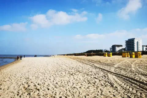 Ein weiter, sonniger Duhnen Strand mit gelben Strandkörben und einigen Spaziergängern. Im Hintergrund moderne Gebäude.