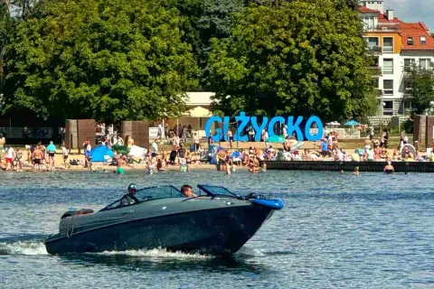 Łódź motorowa płynie po jeziorze, w tle plaża z ludźmi i napis "GIŻYCKO". Idealne miejsce, gdzie na Mazury!