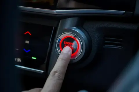 Un doigt appuie sur le bouton "ENGINE START" qui clignote en rouge. La voiture est prête à démarrer.