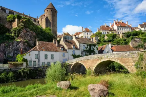 Un village pittoresque de la Bourgogne avec un pont de pierre ancien, des maisons aux toits rouges et une tour médiévale perchée sur une colline verdoyante.