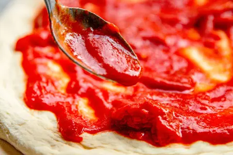 Ein Löffel verteilt die leuchtend rote San Marzano Pizza Sauce auf einem frischen Pizzateig.