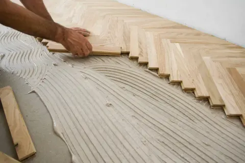 Mains d'un artisan en train de poser du parquet en chevrons sur un lit de colle ondulée.