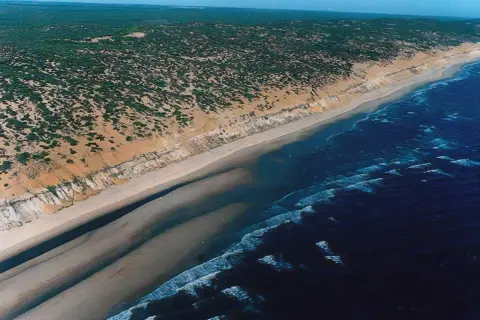 Vista aérea de la **playa de Castilla**, con dunas cubiertas de vegetación, arena dorada y el mar azul con olas.