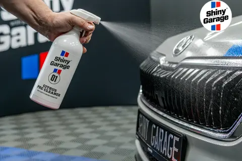 Ręka rozpyla pre-wash oprysk Citrus Pre Cleaner z butelki Shiny Garage na grill samochodu, usuwając brud.