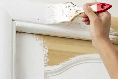 Une main applique de la peinture blanche sur une porte intérieure en bois. Le travail de peinture est en cours, révélant le bois naturel sous la couche blanche.