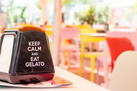 Na stoliku stoi czarny dozownik na serwetki z napisem "Keep calm and eat gelato". W tle kolorowe krzesła w kawiarni. To dobre hasło na lato!