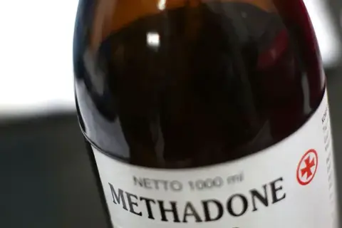 Butelka z ciemnobrązowym płynem, na etykiecie napis "Methadone" i czerwony symbol krzyża.