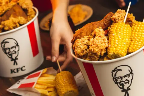 Wiadro KFC z kurczakiem, frytkami i kolbami kukurydzy.