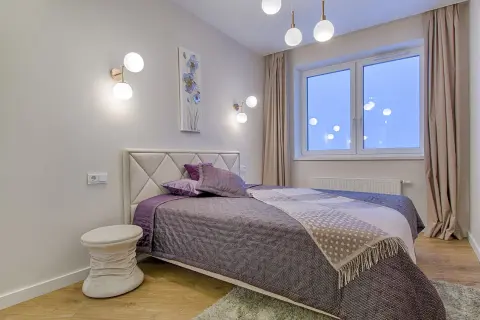 Une chambre moderne aux murs clairs, prête à peindre. Un lit douillet avec des coussins violets et une couverture à motifs. Des appliques murales design et une fenêtre lumineuse.