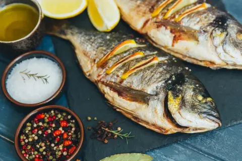 Zwei gebratene Dorade Fische mit Zitronenscheiben, Olivenöl, Salz und Pfefferkörnern auf einem dunklen Brett.