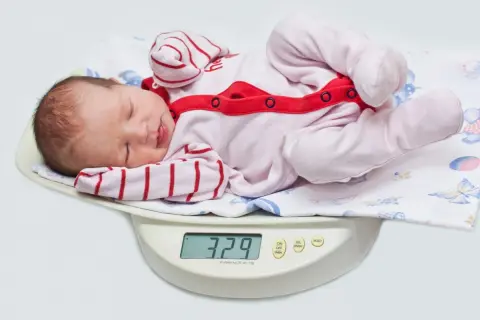 Nowe życie waży 3,29 kg. Dziecko po urodzeniu leży na wadze, ubrane w różowy pajacyk w paski.