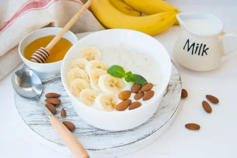 Pyszna owsianka proteinowa z bananem, migdałami i miętą. Obok miseczka z miodem i dzbanuszek z mlekiem.