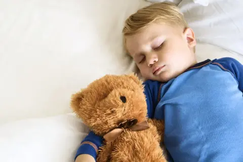 En liten pojke sover sött med sin nallebjörn. Hur många timmar ska en 10-åring sova? Sömn är viktigt för barn.