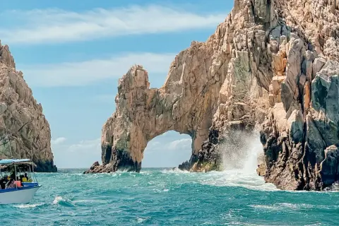 Un paseo en barco revela el Arco de Cabo San Lucas, una maravilla natural que ver en Baja California. Las olas rompen contra las rocas doradas bajo un cielo azul.
