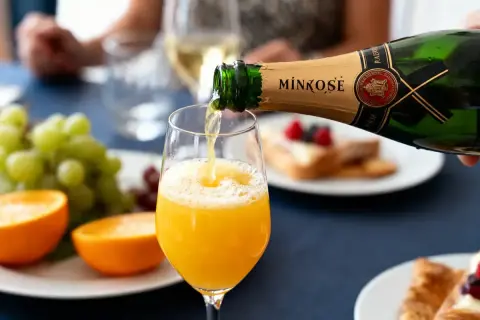 Une bouteille de Mimosé verse son contenu pétillant dans un verre rempli de jus d'orange. Un brunch festif se prépare.