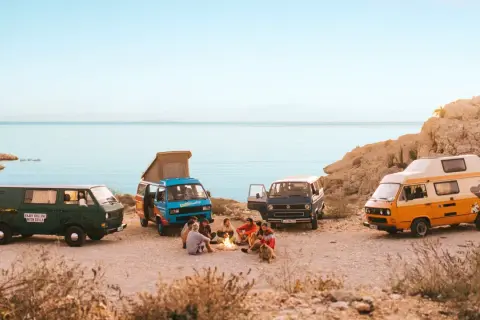Vier bunte Campervans am Meer, Menschen sitzen am Lagerfeuer.
